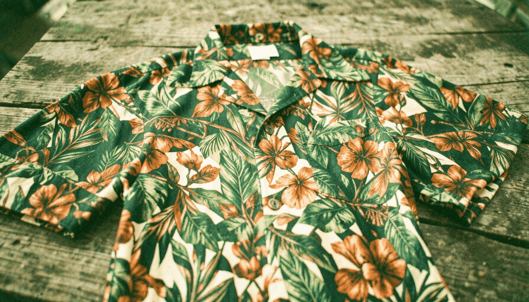 A warm, sunlit photograph of a richly patterned Hawaiian shirt with tropical leaf and floral motifs in deep greens, burnt orange, and cream, laid across weathered wood with visible film grain.