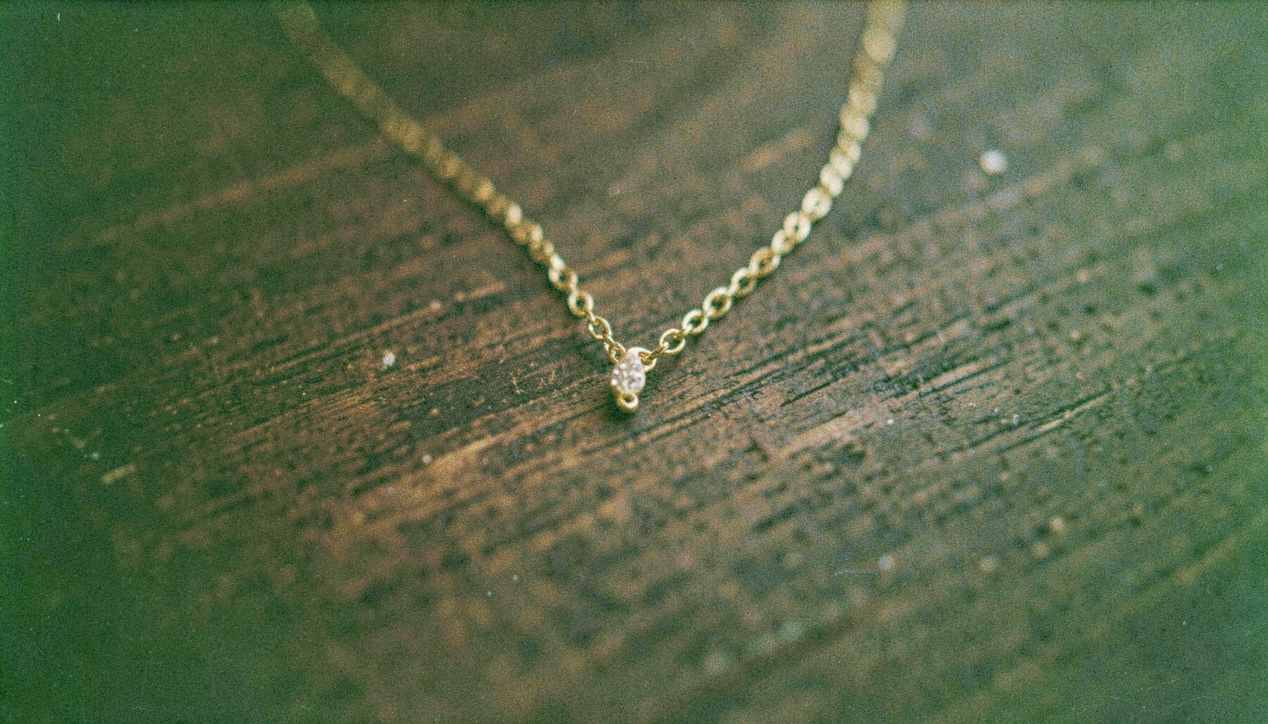 An analog film close-up of a delicate pendant necklace on dark wood, subtle highlights on the chain, deep browns and soft gold.