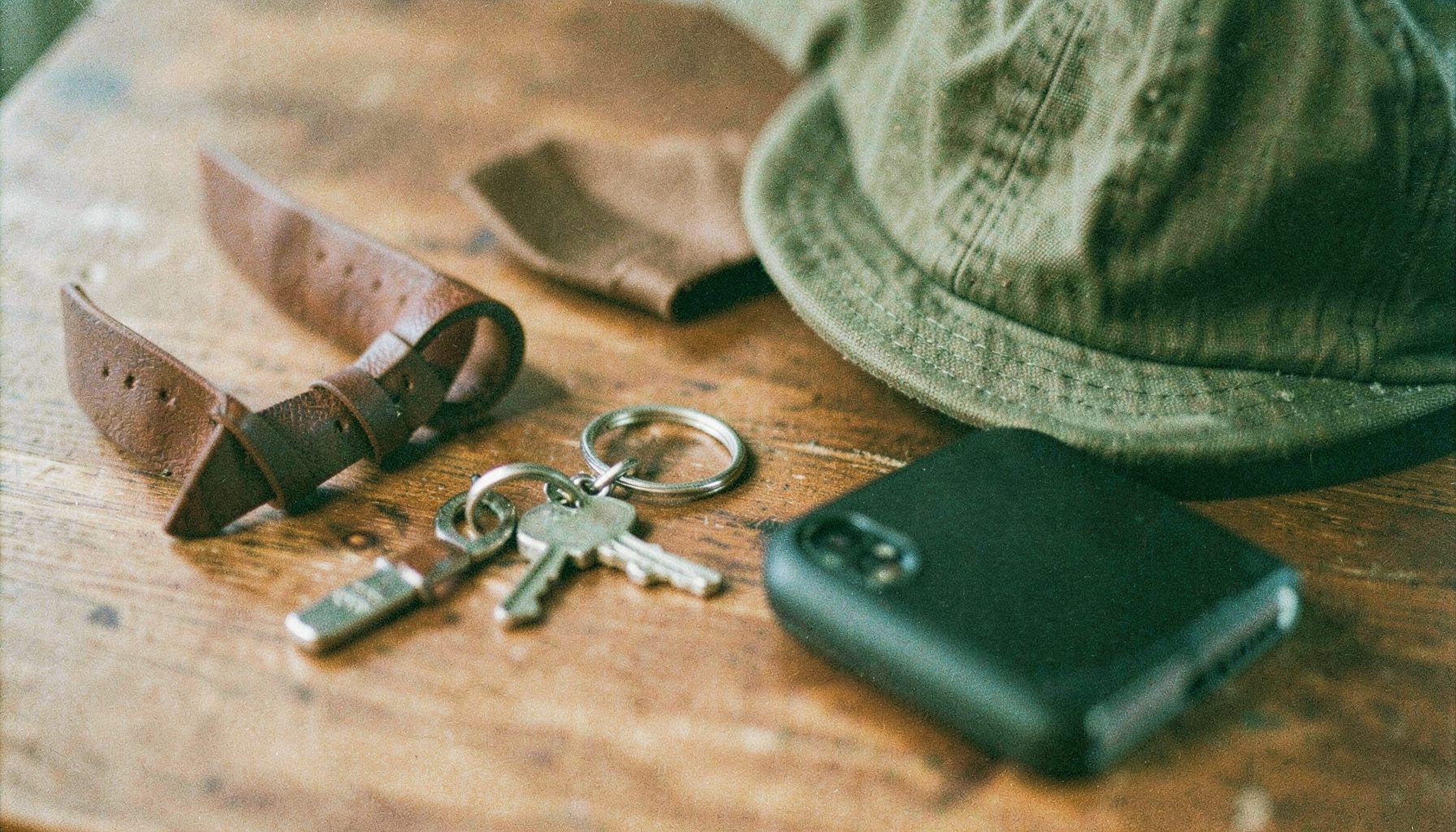 A close-up analog film photograph of assorted personal accessories laid on a warm wooden surface — a leather watch band, a metal keychain, a canvas cap, and a phone case, with soft natural light and visible film grain, rich browns and muted greens.