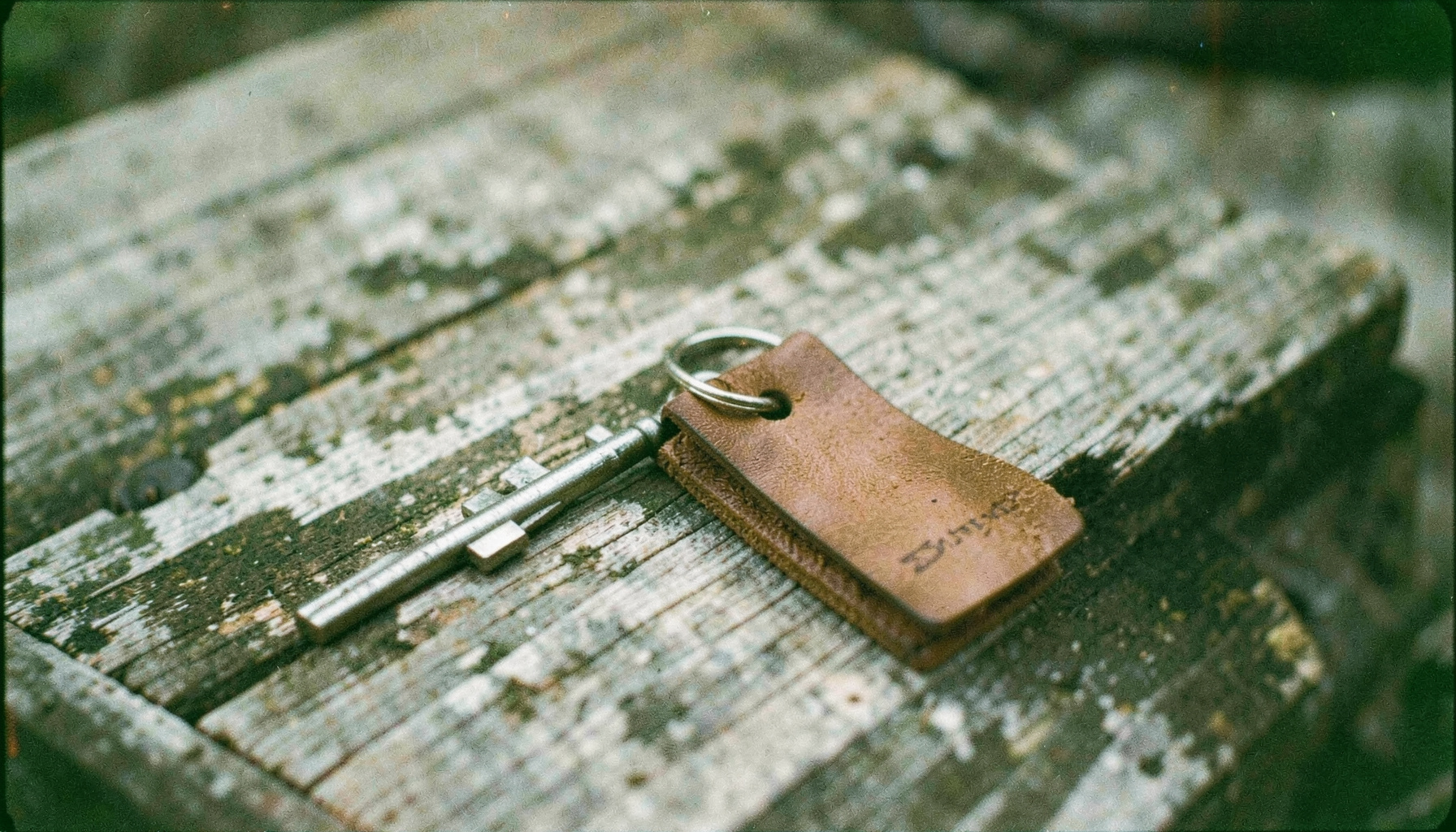 An analog film photograph of a metal keychain with a leather tag, sitting on a weathered wooden bench, muted grays and warm browns.