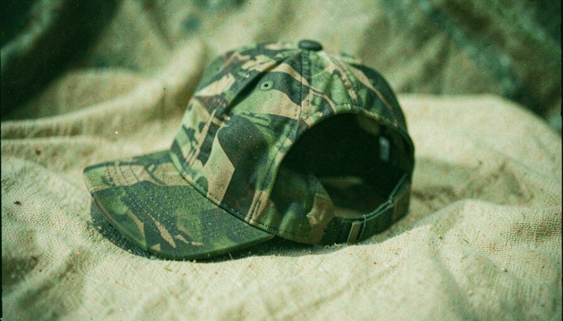 An analog film close-up of a camo-print ponytail baseball cap with an open back, resting on natural canvas fabric, earthy greens and tans.