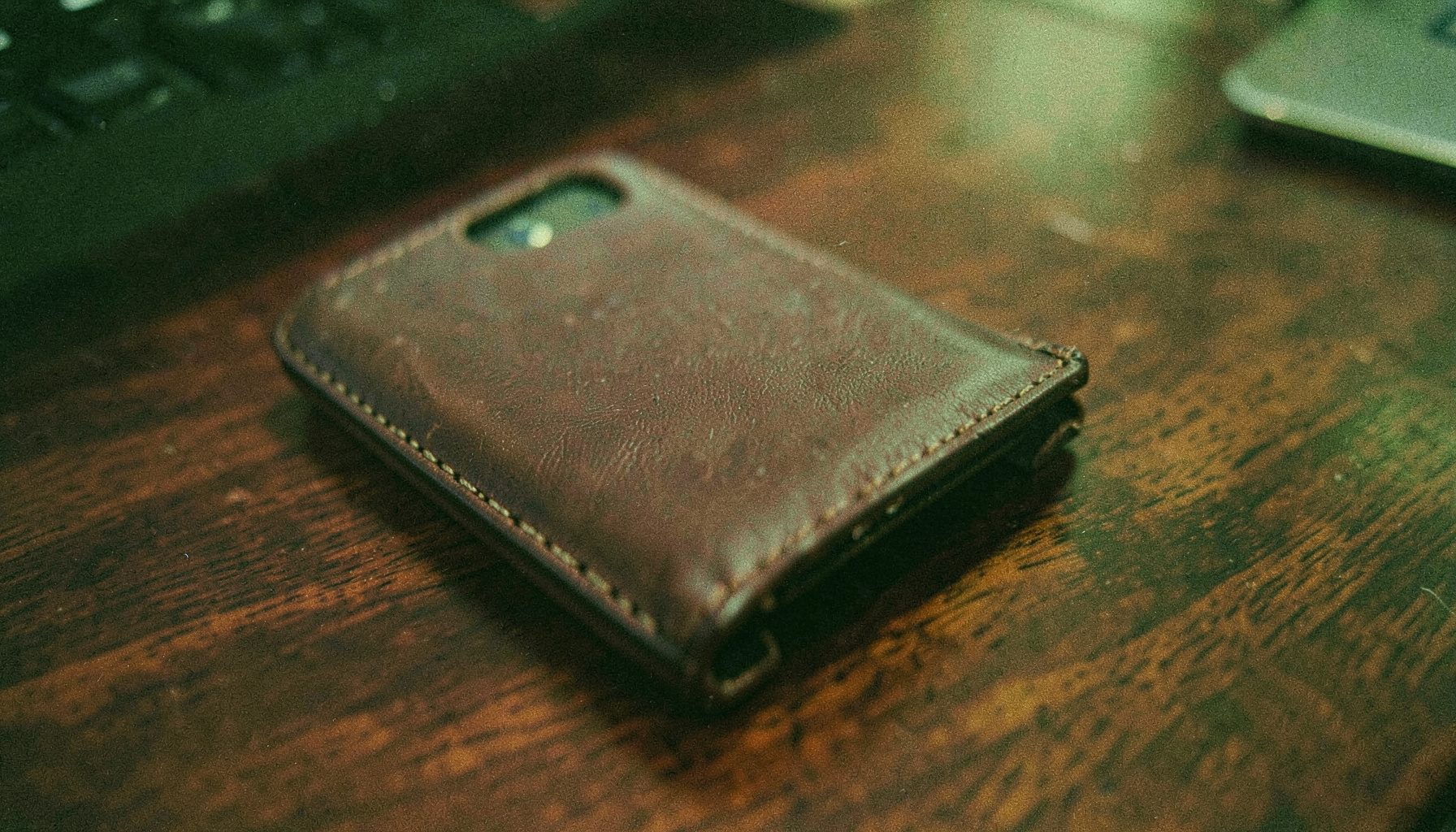 An analog film close-up of a leather phone case with visible stitching, resting on a dark walnut desk, rich browns and warm highlights.