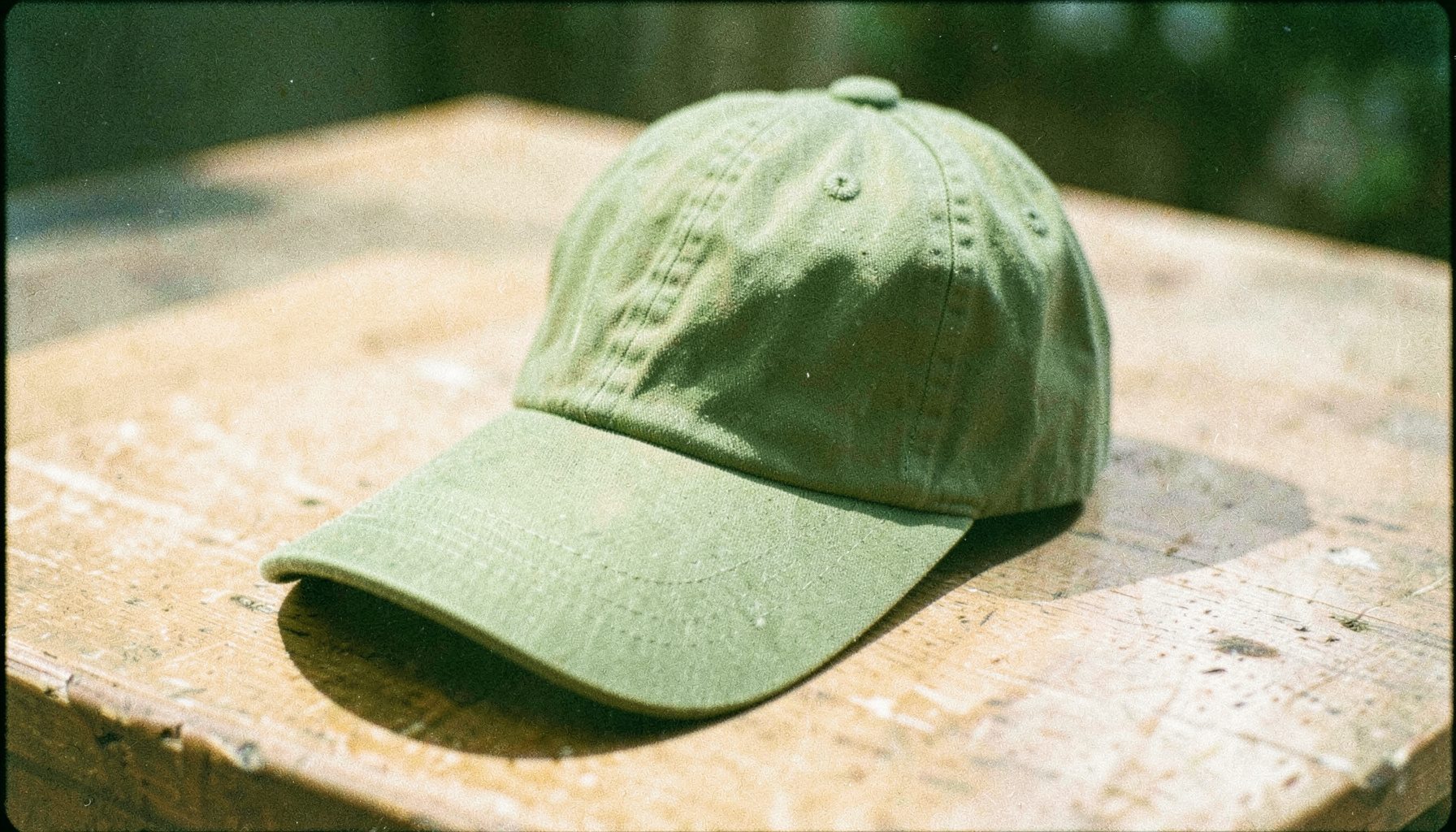 An analog film photograph of a classic baseball cap in olive green on a sunlit wooden table, soft shadows and visible film grain.