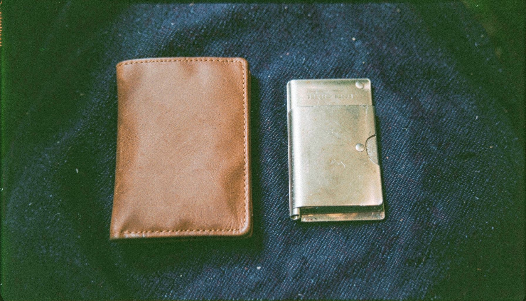 An analog film photograph of a slim leather wallet and a metal card holder side by side on dark denim, warm browns and indigo.