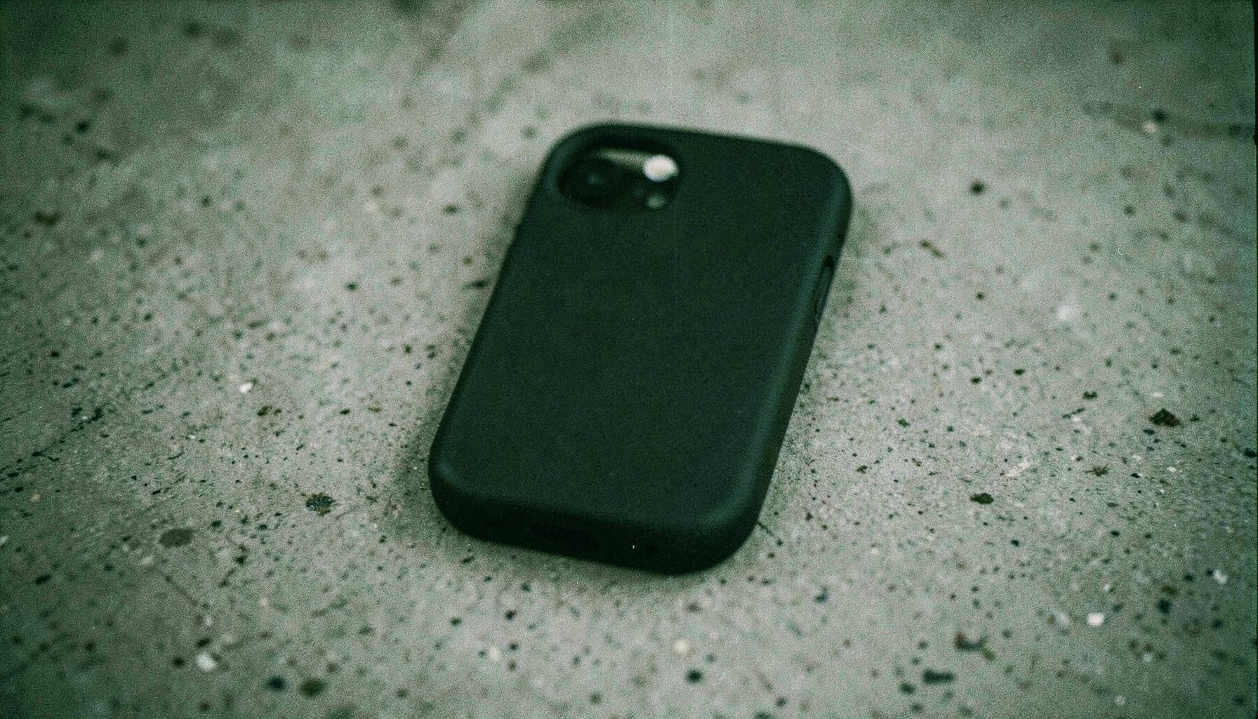 An analog film photograph of a rigid phone case in matte black on a concrete surface, side lighting, moody grays and deep blacks.