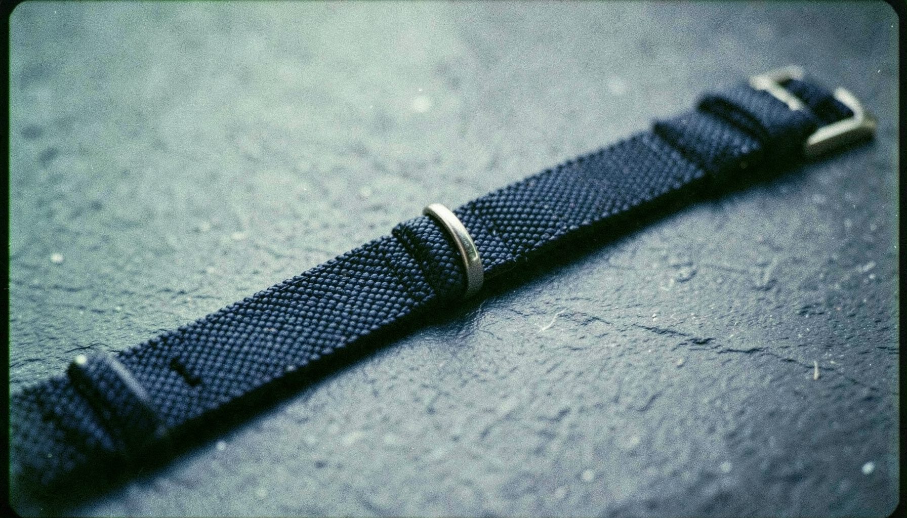 An analog film close-up of a woven watch band unfastened on a slate surface, subtle texture, cool grays and deep navy.