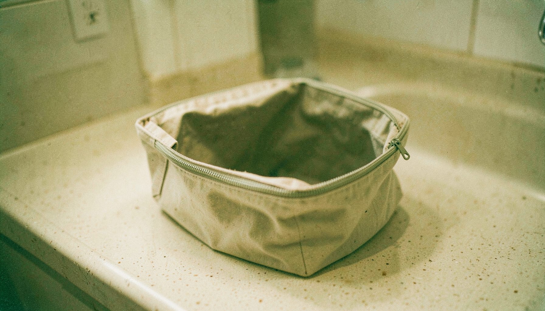 An analog film photograph of an open canvas toiletry bag with a zipper, placed on a bathroom counter, soft warm light, beige and olive tones.